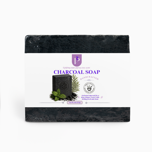 A packaged bar of charcoal soap centered on a light gray background. The soap is wrapped in a white paper label with purple text reading 'CHARCOAL SOAP' and a small product photo of a dark soap bar with mint leaves and a rosemary sprig.