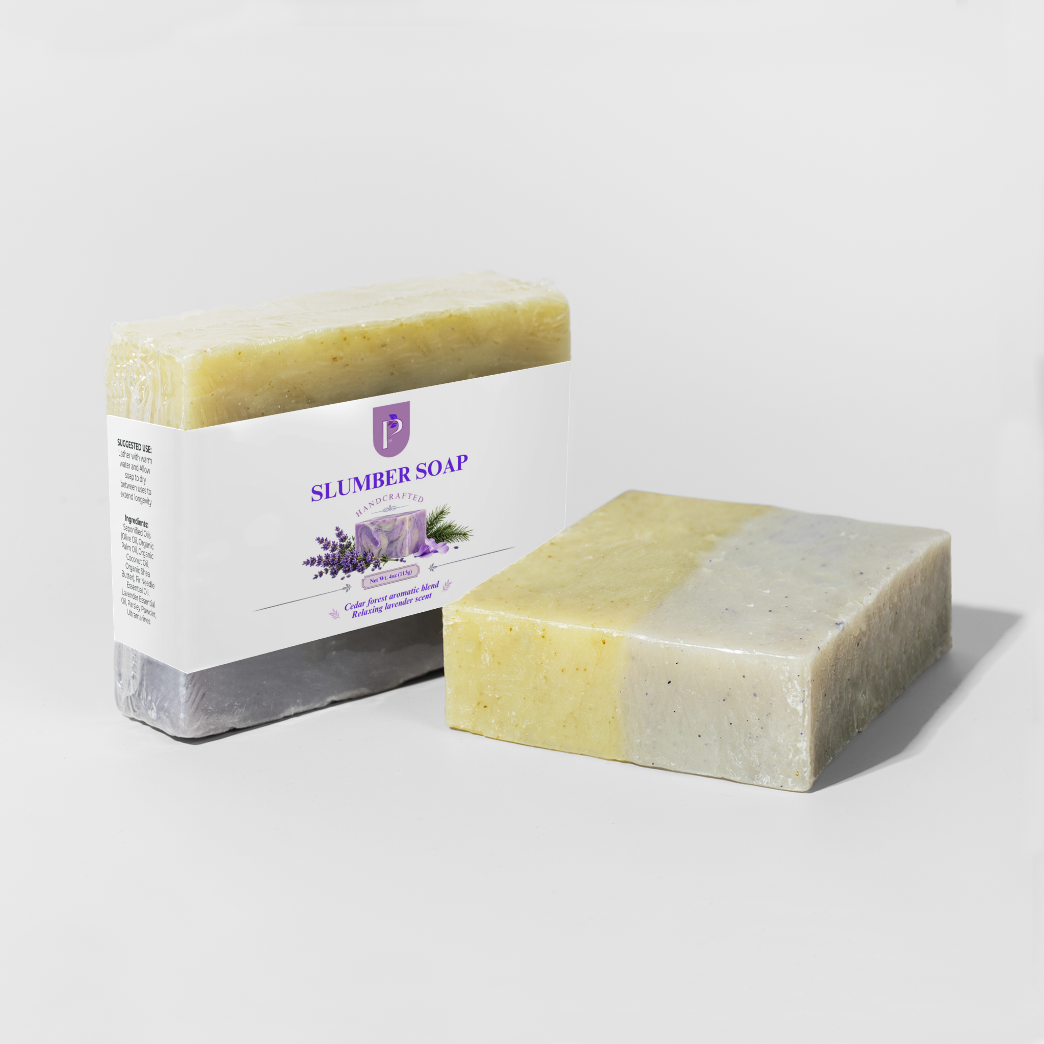 A bar of Slumber Soap shown next to its paper packaging box on a light gray studio background. The soap bar is two-toned with a pale yellow upper half and a light gray lower half, while the box is white with purple branding and lavender illustrations.