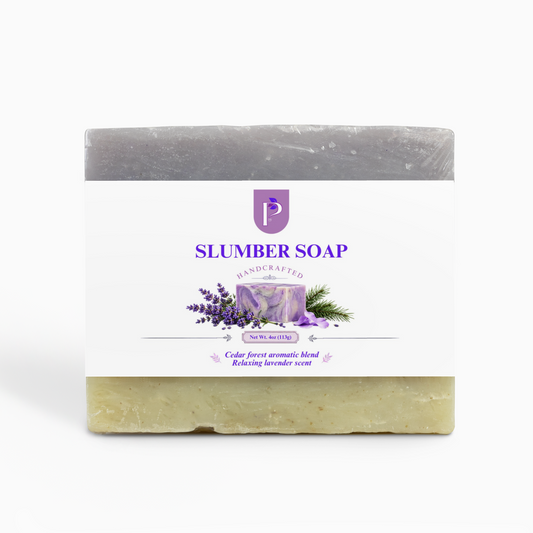 A rectangular bar of soap with a white paper label reading 'SLUMBER SOAP' and 'Handcrafted'. The top half of the soap is gray, and the bottom half is a pale yellow-green with visible botanical flecks.