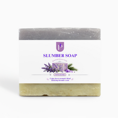 A rectangular bar of soap with a white paper label reading 'SLUMBER SOAP' and 'Handcrafted'. The top half of the soap is gray, and the bottom half is a pale yellow-green with visible botanical flecks.