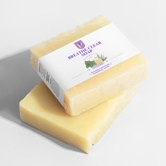 Two rectangular bars of Breathe Clear Soap stacked on a white surface, with one bar resting on top and wrapped in a white paper label. The soap is pale yellow, and the label features purple branding and a small lemon and green herb illustration.