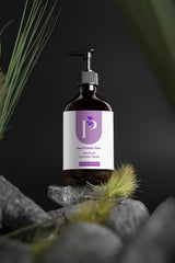 A pump bottle of hand and body wash with a white and purple label, styled on gray stones with green grass against a dark background.