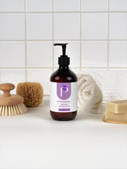 A pump bottle of hand and body wash with a purple and white label, styled on a white surface with a rolled towel, soap bar, sponge, and brush against a white tiled background.