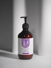 An amber glass pump bottle of hand and body wash with a black dispenser and a white label featuring a purple logo, set against a gray background with vertical ribbed panels.