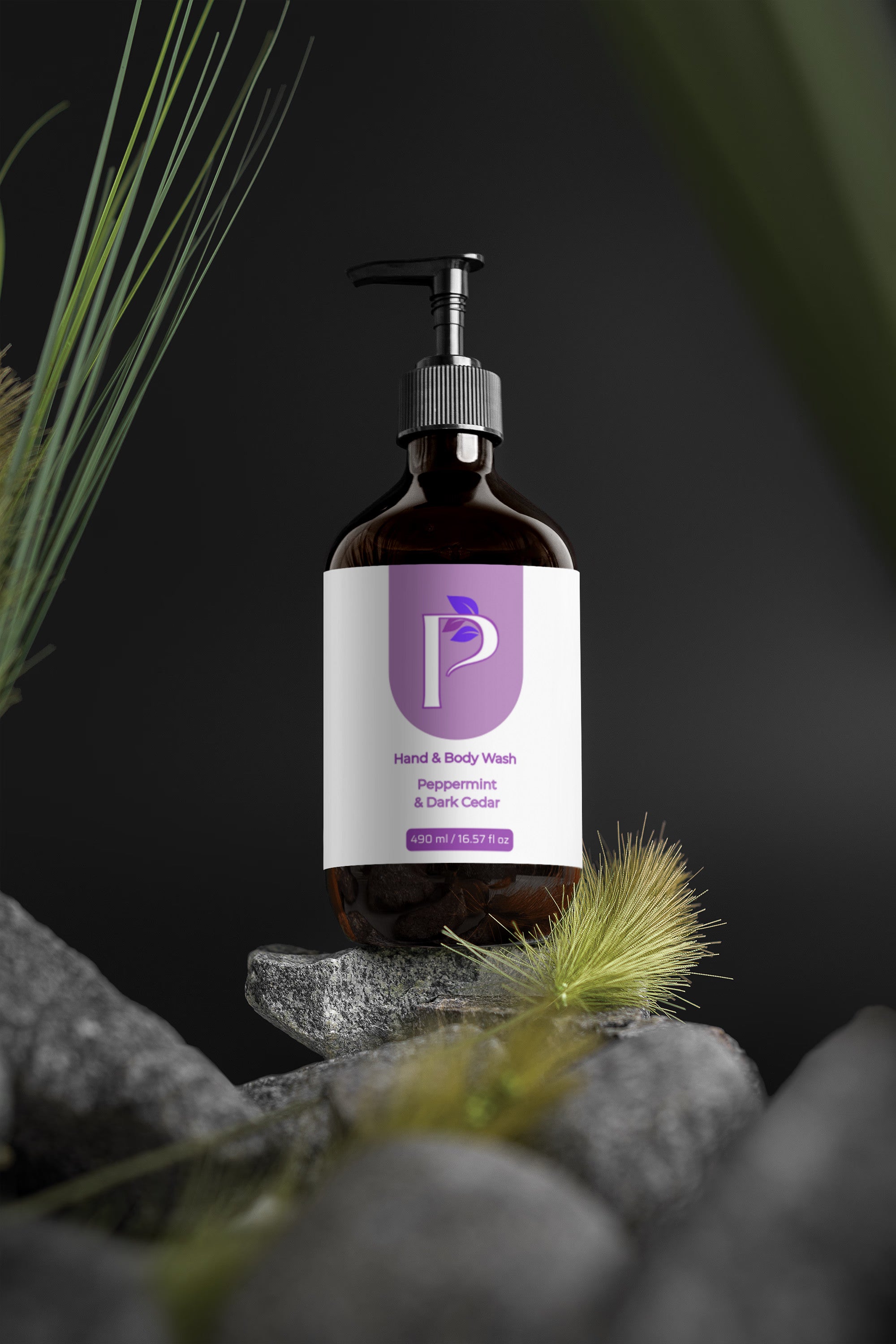 A dark amber pump bottle of hand and body wash with a white and purple label, styled on gray stones with dried grass against a dark background.