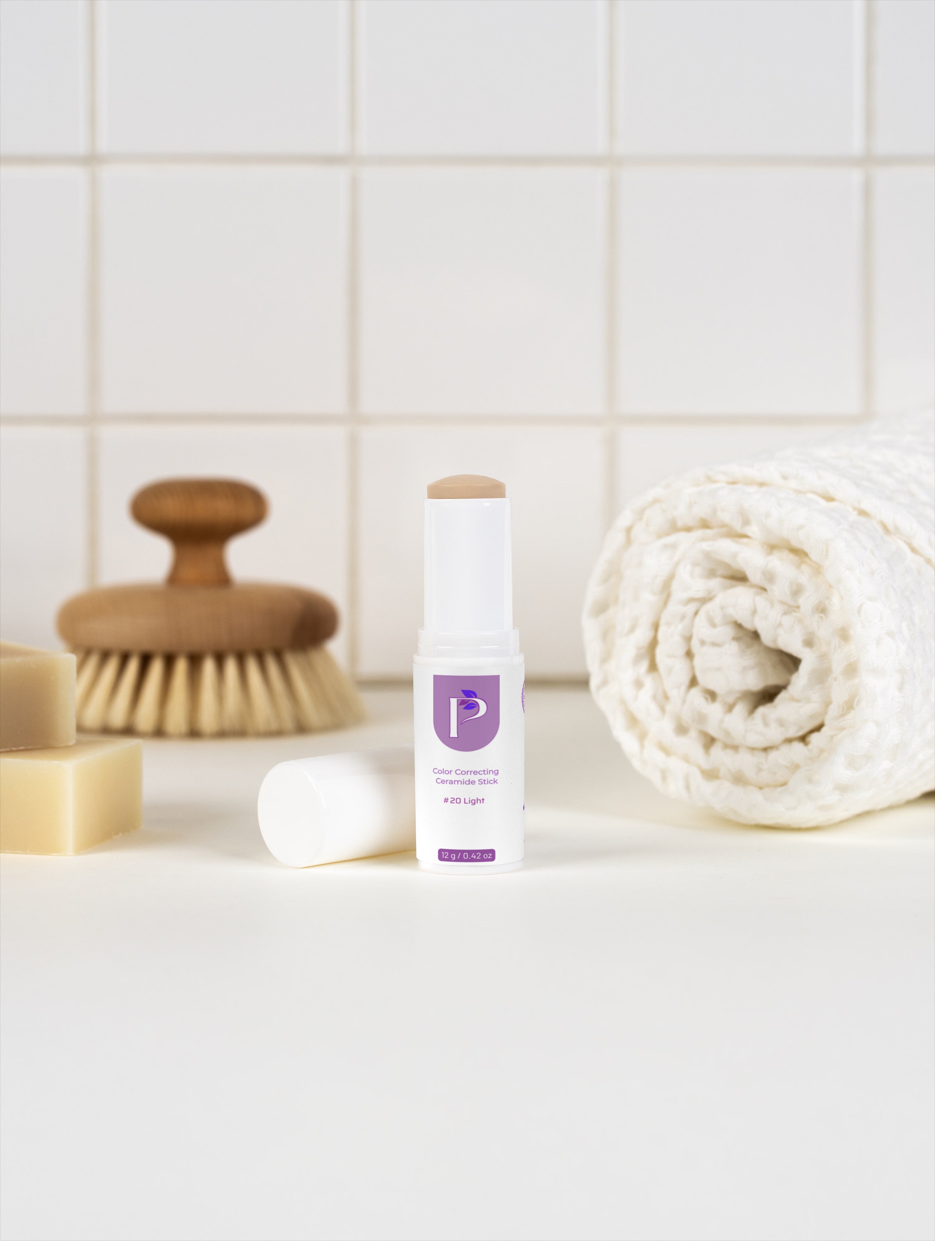 A white and purple cosmetic stick product stands on a white surface with its cap removed beside it. The scene includes a rolled cream towel, a wooden-handled brush, soap bars, and a tiled wall background.