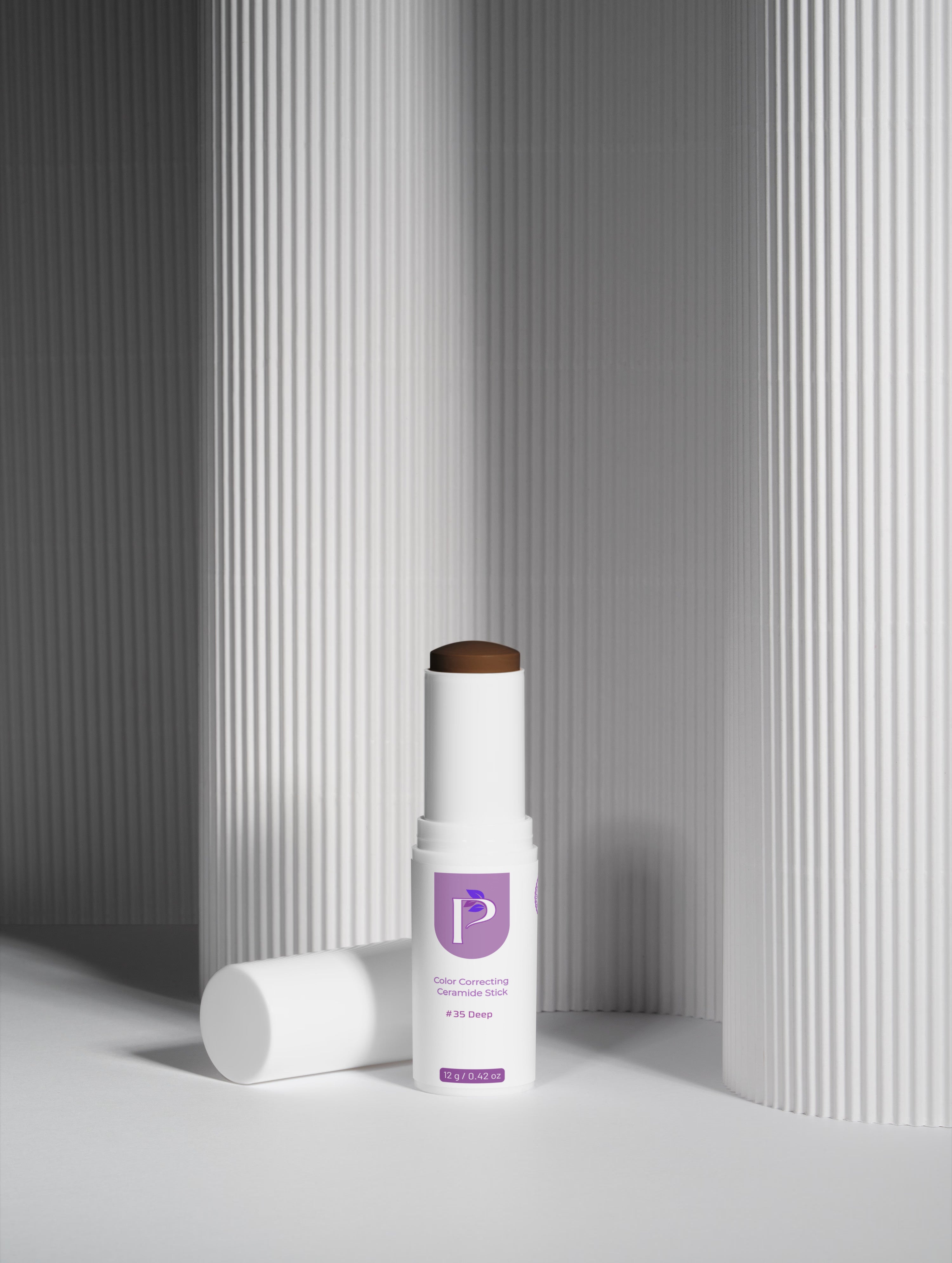 A white cosmetic stick container with a purple logo label stands upright on a white surface, with its matching cap lying beside it. The product appears to be a skin-brightening or complexion stick, photographed in a clean studio setup with fluted gray columns in the background.