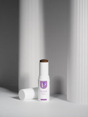 A white cosmetic stick container with a purple logo label stands upright on a white surface, with its matching cap lying beside it. The product appears to be a skin-brightening or complexion stick, photographed in a clean studio setup with fluted gray columns in the background.