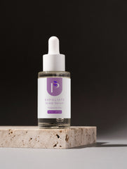 A clear glass dropper bottle of Exfoliate Scalp Serum with a white label and purple logo, displayed on a textured stone block against a dark gray background.