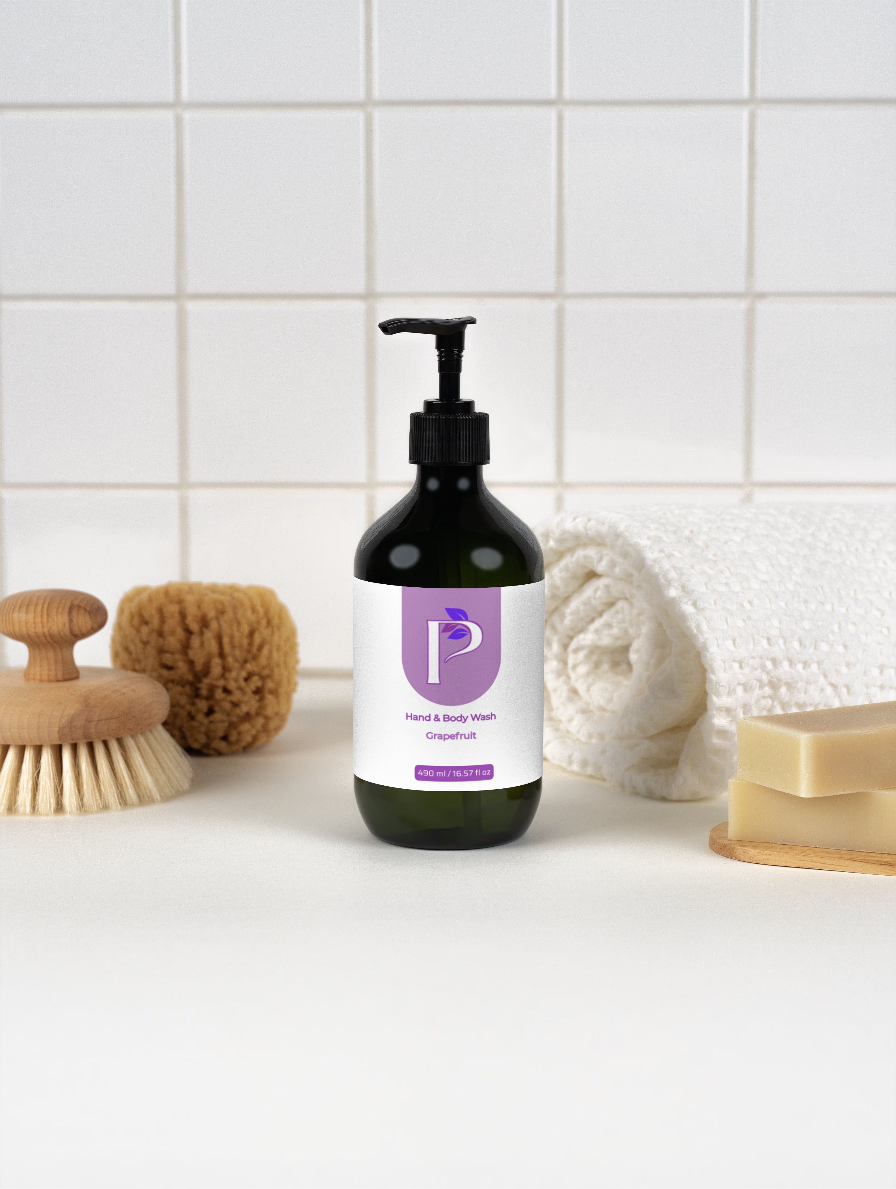 A dark green pump bottle of hand and body wash with a purple and white label, styled on a white surface with a wooden brush, sponge, rolled towel, and soap bar against a white tiled background.