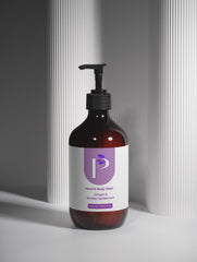 An amber glass pump bottle of hand and body wash with a black dispenser and a white label featuring a purple logo, set against a gray background with vertical ribbed panels.