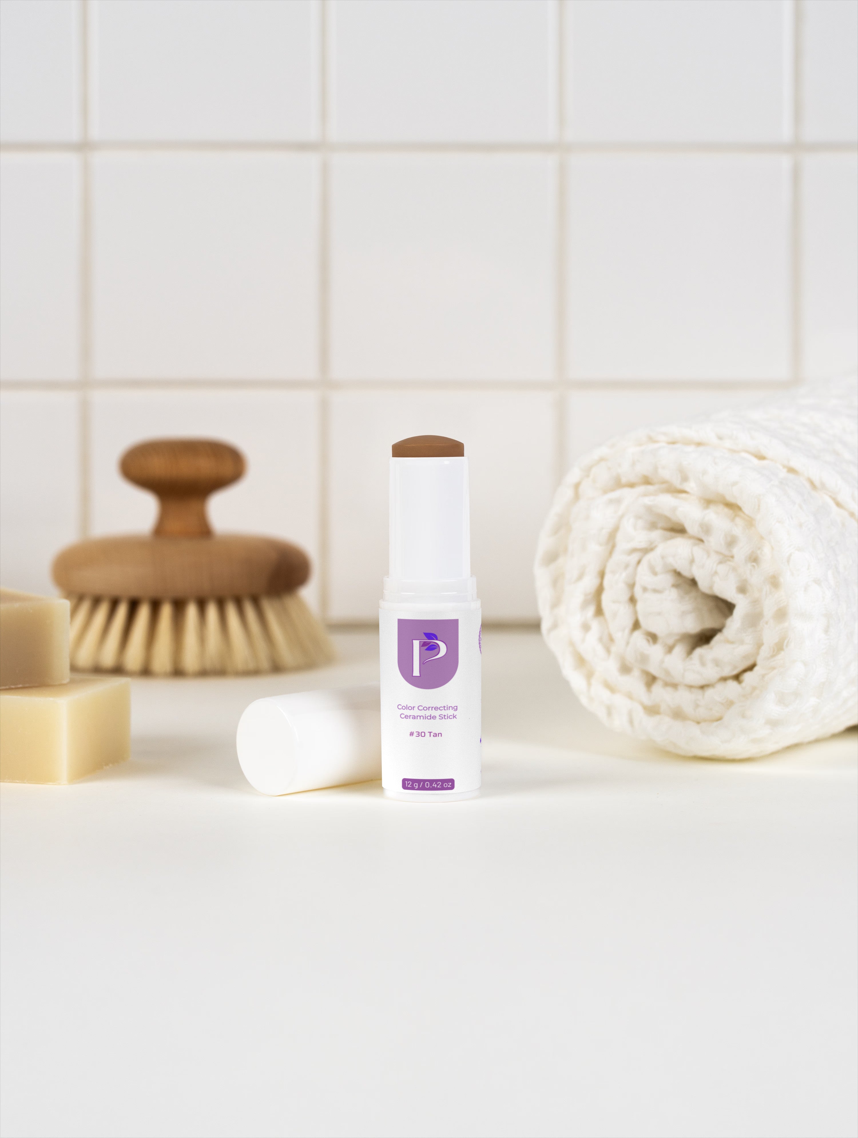 A white and purple cosmetic stick product stands on a white surface with its cap removed beside it. The scene includes a rolled cream towel, a wooden-handled brush, soap bars, and a tiled wall in the background.