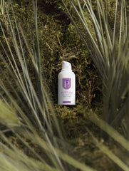 A small white skincare bottle labeled 'PEPTIDE REPAIR CREAM' with a purple logo, placed on dark moss surrounded by tall grass blades.