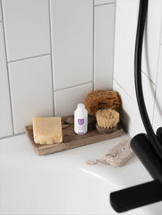 Bathroom corner with soap, bottle, and scrubber on a wooden tray against a tiled wall.