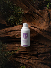 A white pump bottle of Peptide Eye Cleanser with a purple label, placed on a textured brown log with green pine needles in the background.