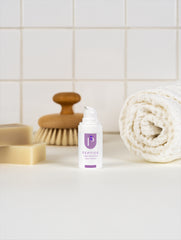 A small white pump bottle labeled 'PEPTIDE' with a purple logo, standing on a white surface in front of a tiled wall. A rolled cream towel, a wooden-handled brush, and soap bars are placed around it as styled props.